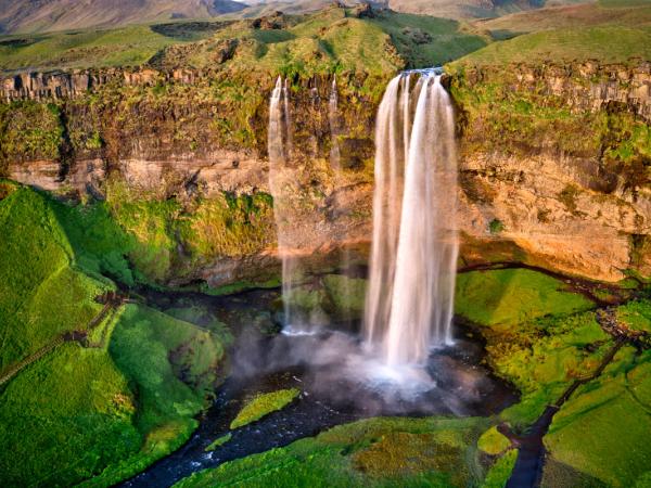 A grand waterfall cascades over a green, rocky cliff into a river, surrounded by vibrant green hills and mountains.