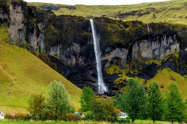 A tall waterfall cascades down a dark, mossy cliff flanked by green hills and trees, with a small red-roofed house visible on the left.