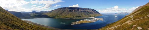 Panoramic view of a coastal town on a peninsula in a fjord, surrounded by steep mountains, with a ship in the water.