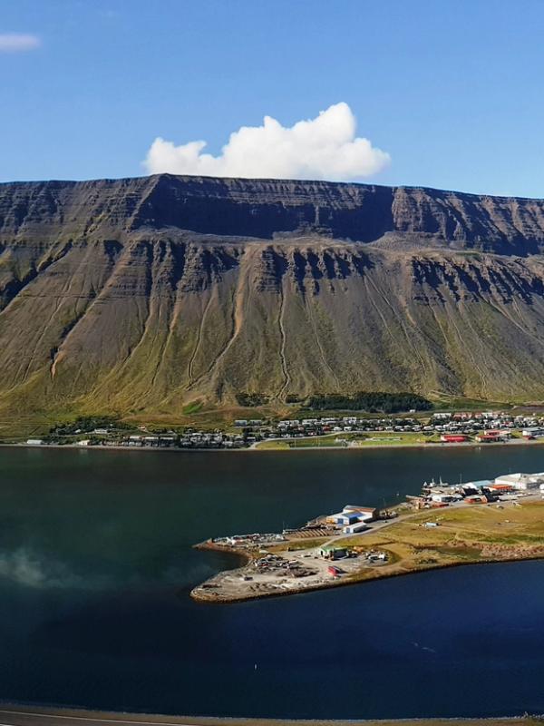 Panoramic view of a coastal town on a peninsula in a fjord, surrounded by steep mountains, with a ship in the water.