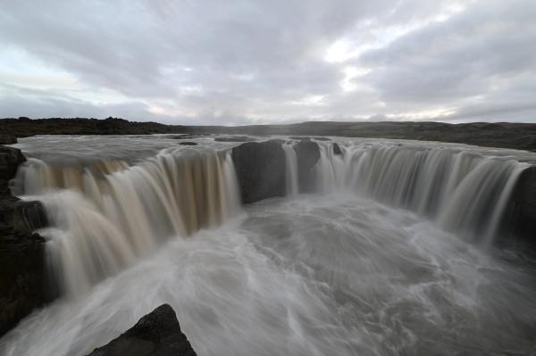 Wide, powerful waterfall with milky, blurred water cascading over dark rocks under a cloudy sky.