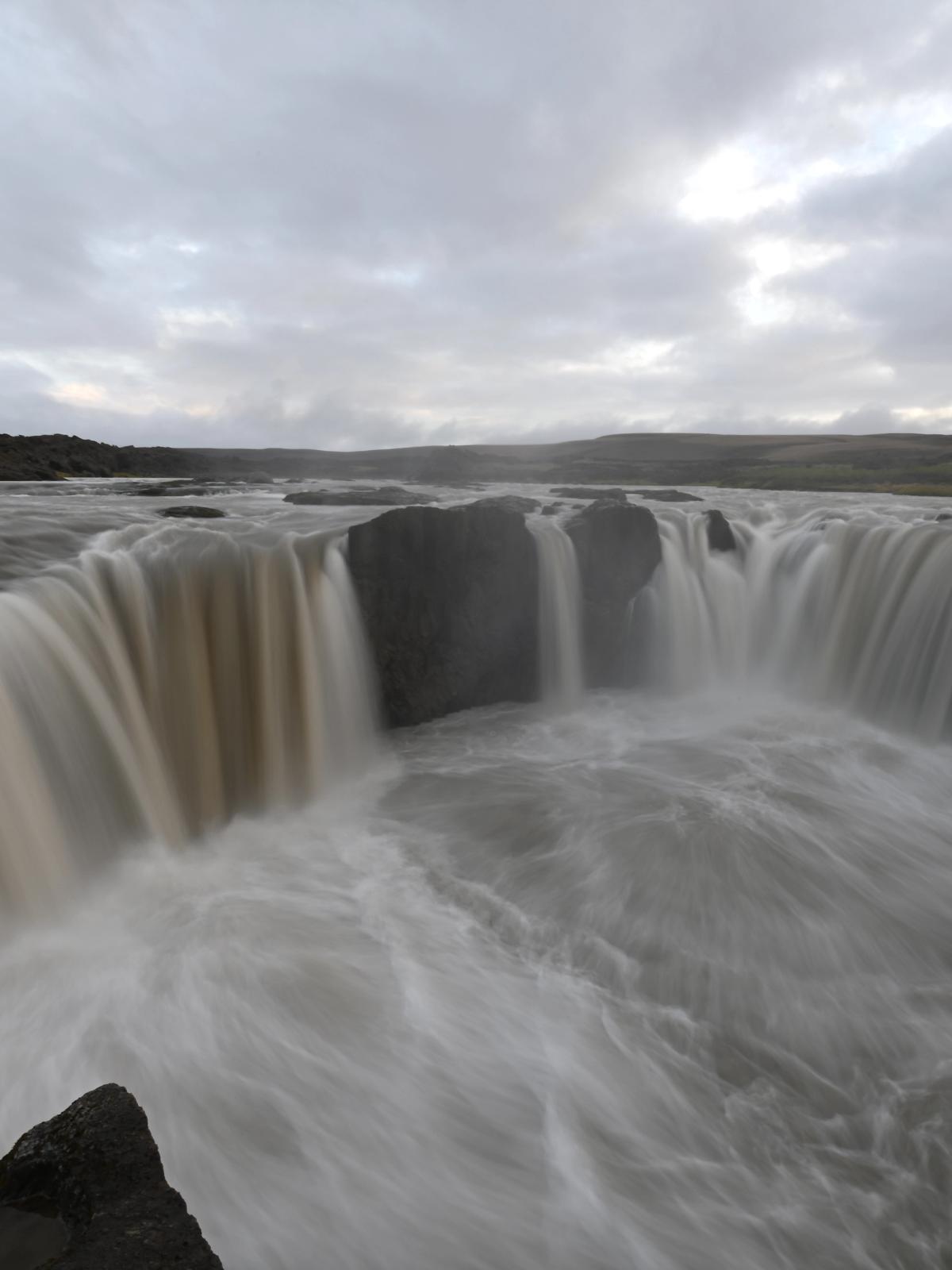 Wide, powerful waterfall with milky, blurred water cascading over dark rocks under a cloudy sky.