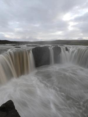 Wide, powerful waterfall with milky, blurred water cascading over dark rocks under a cloudy sky.