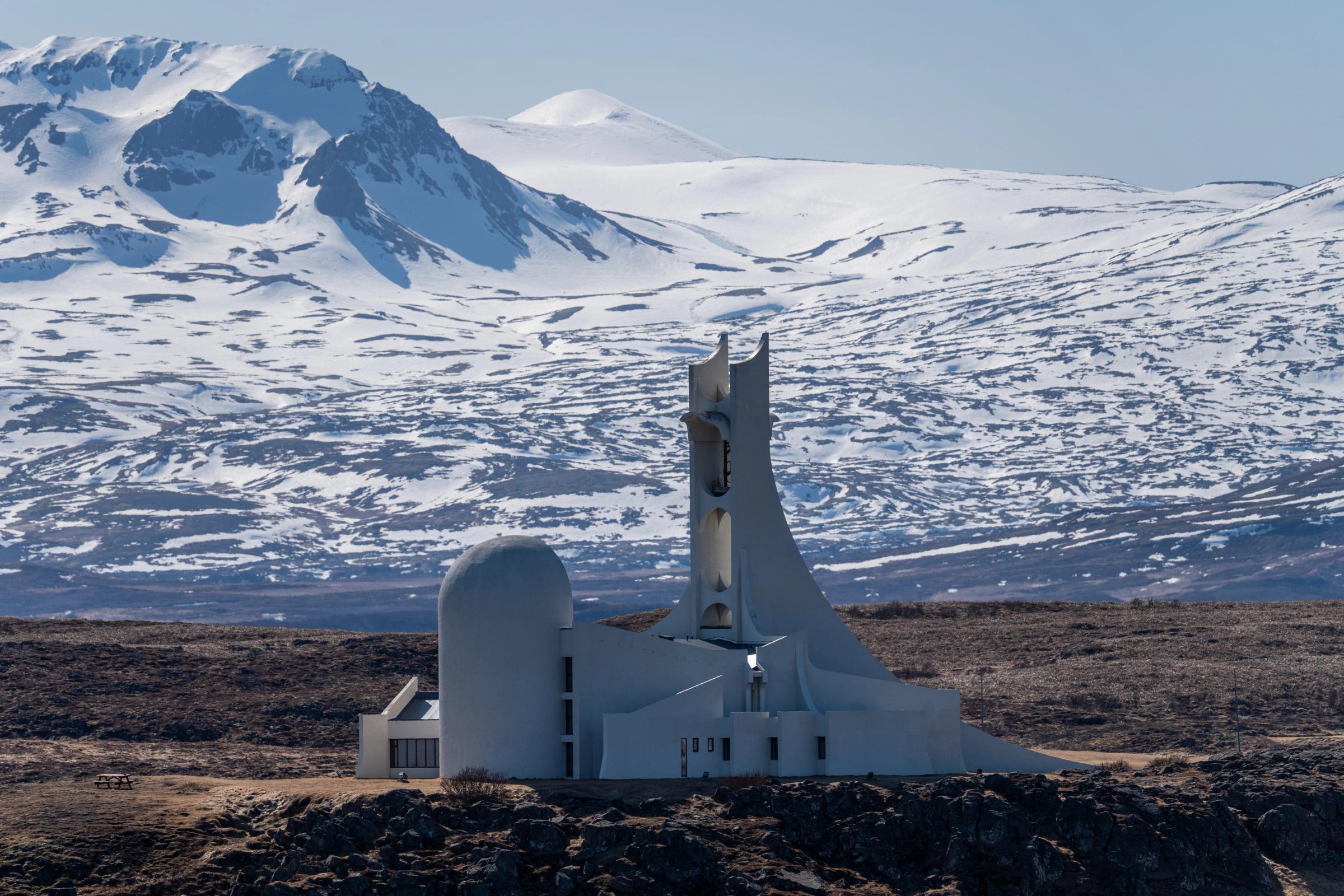 A striking white modernist church with a tall, angular bell tower stands on a rocky plain, backed by snow-covered mountains.