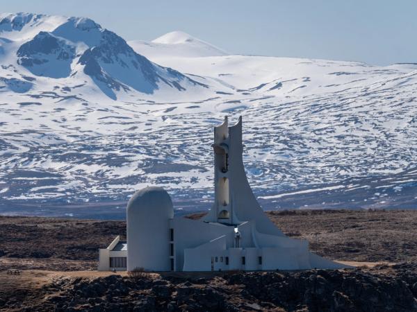 A striking white modernist church with a tall, angular bell tower stands on a rocky plain, backed by snow-covered mountains.