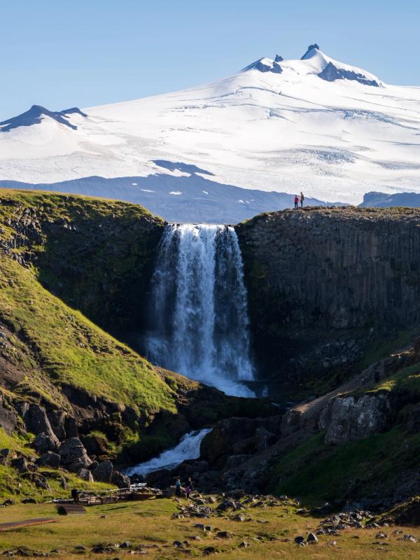 A waterfall cascades over a basalt cliff with two small figures, backed by a large snow-capped mountain.