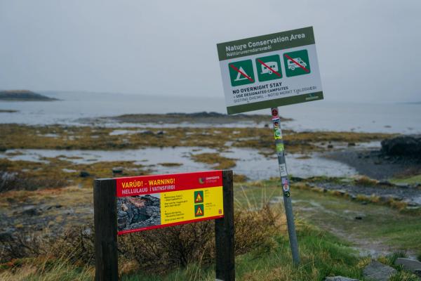 Two warning signs, one prohibiting overnight stays and another cautioning about dangerous ground, stand by a cloudy bay.