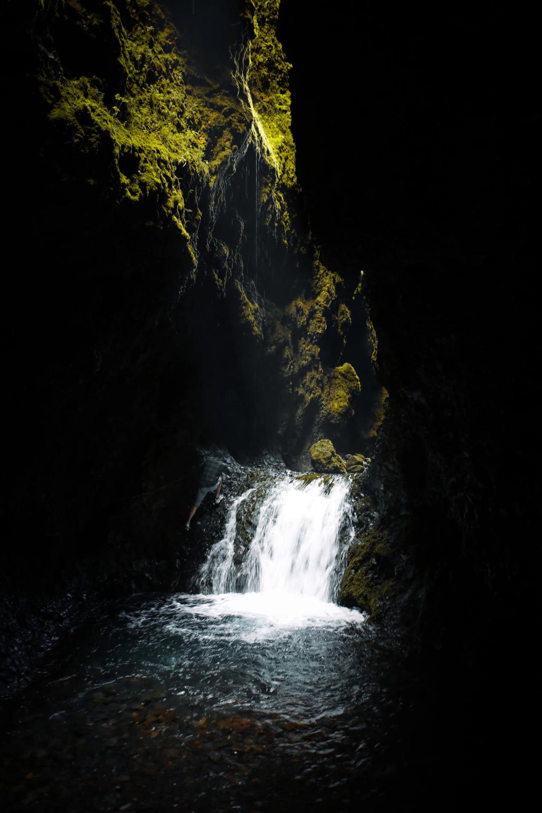 Una cascada se precipita en una oscura poza dentro de una cueva, iluminada por la luz que resalta el musgo verde vibrante en las paredes rocosas.