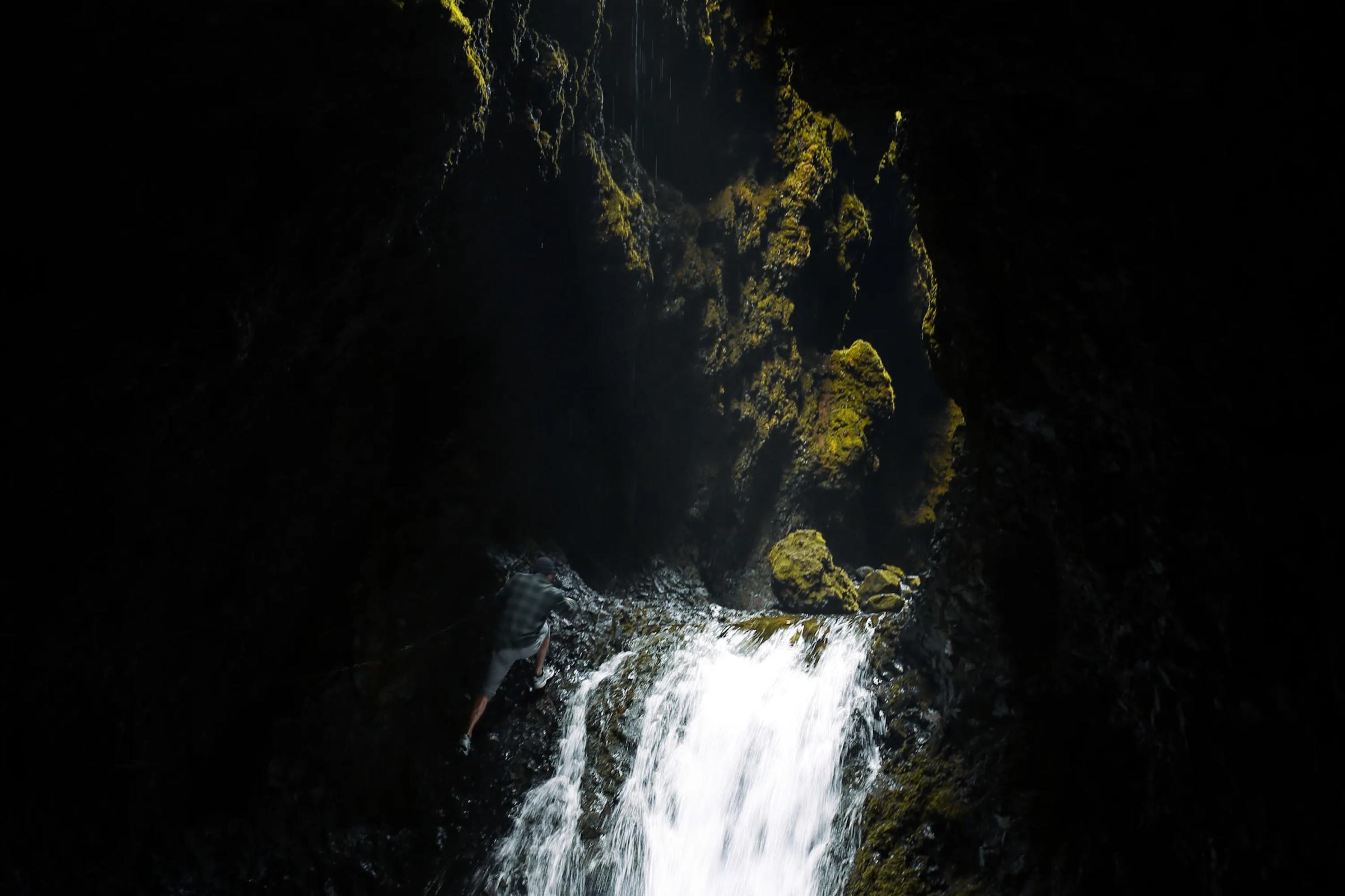 Una cascada se precipita en una oscura poza dentro de una cueva, iluminada por la luz que resalta el musgo verde vibrante en las paredes rocosas.