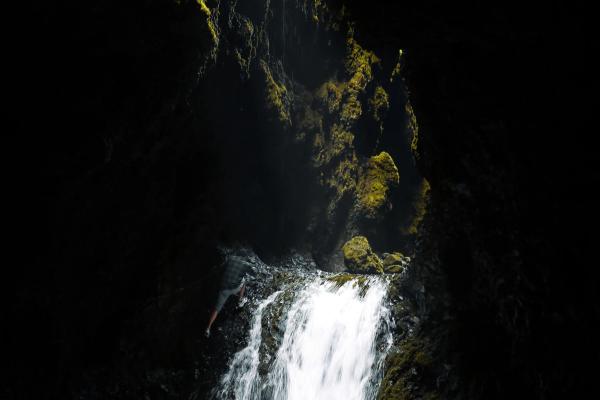 A waterfall cascades into a dark pool inside a cave, illuminated by light highlighting vibrant green moss on the rocky walls.