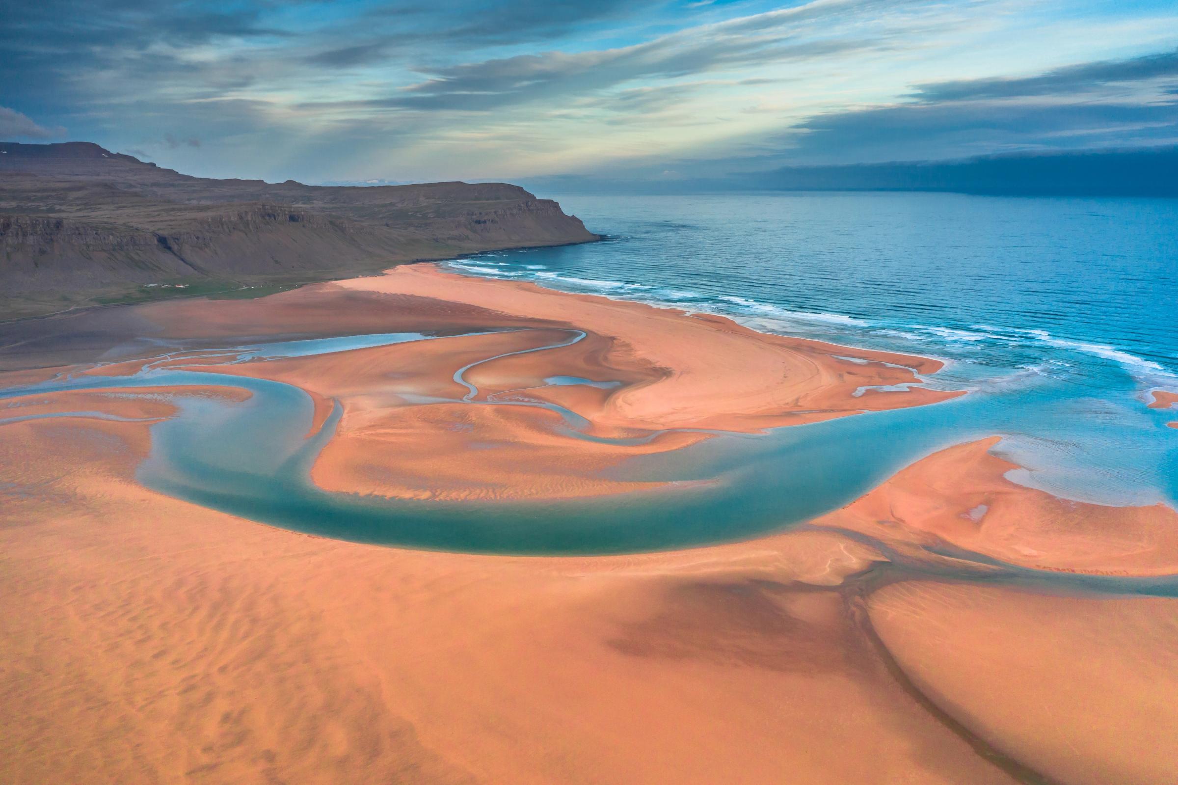 Aerial view of an orange sandy beach with winding blue tidal channels flowing into the ocean, bordered by a rugged coastline under a cloudy sky.