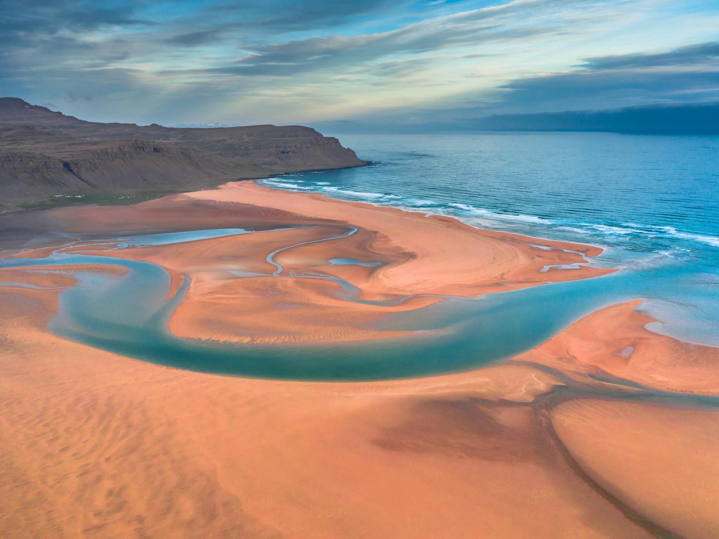 Aerial view of an orange sandy beach with winding blue tidal channels flowing into the ocean, bordered by a rugged coastline under a cloudy sky.
