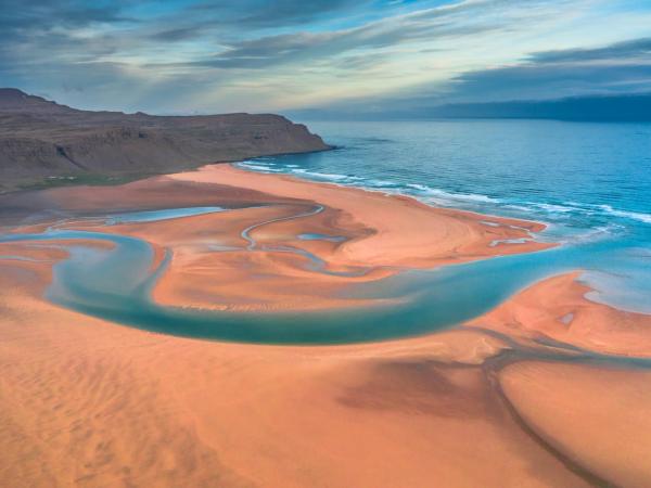 Vista aérea de una playa arenosa de color naranja con sinuosos canales de marea azules desembocando en el océano, bordeada por una costa accidentada bajo un cielo nublado.