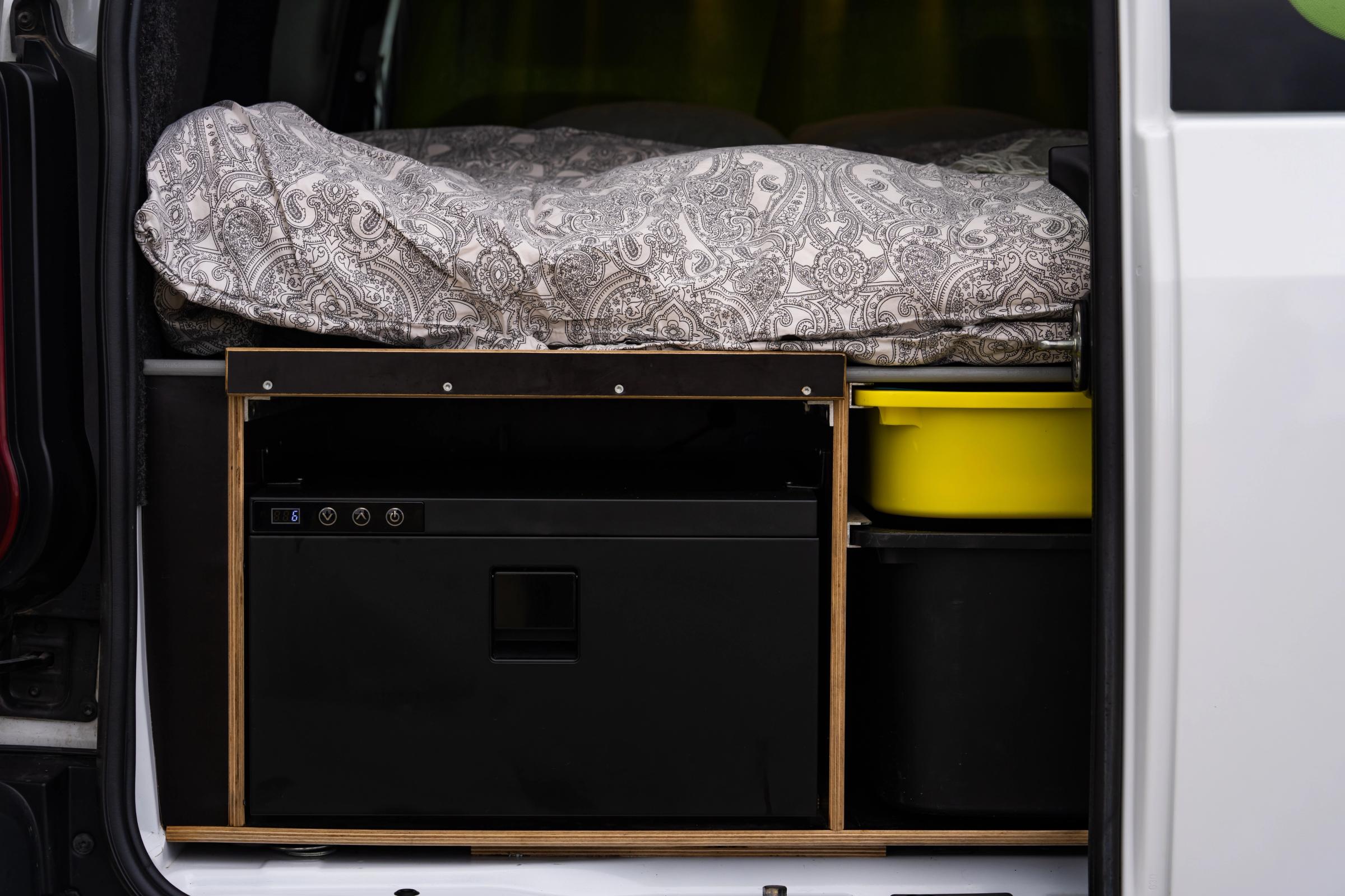 Cajón frigorífico cerrado con botones de control, junto a un recipiente amarillo y un compartimento de almacenamiento debajo de la cama