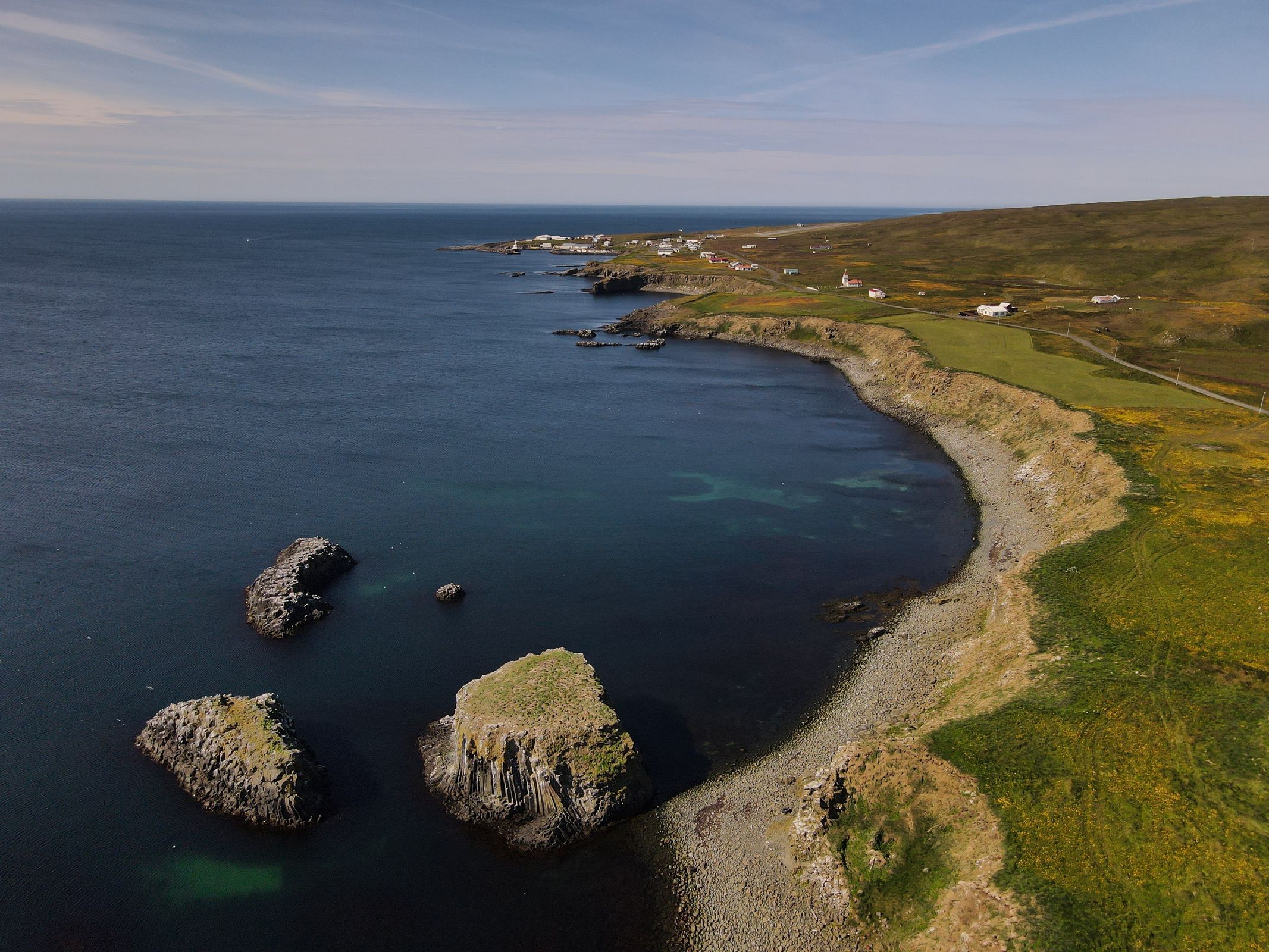Vista aérea de una costa accidentada con un mar azul oscuro, tres grandes pilares de piedra, una playa rocosa curvada y un acantilado cubierto de hierba que conduce a colinas onduladas salpicadas de flores amarillas y un lejano pueblo costero.