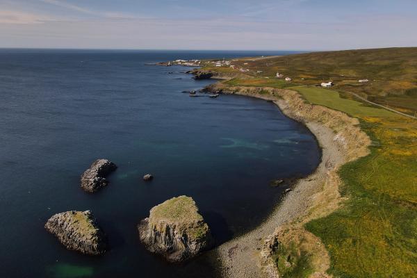 Vista aérea de una costa accidentada con un mar azul oscuro, tres grandes pilares de piedra, una playa rocosa curvada y un acantilado cubierto de hierba que conduce a colinas onduladas salpicadas de flores amarillas y un lejano pueblo costero.