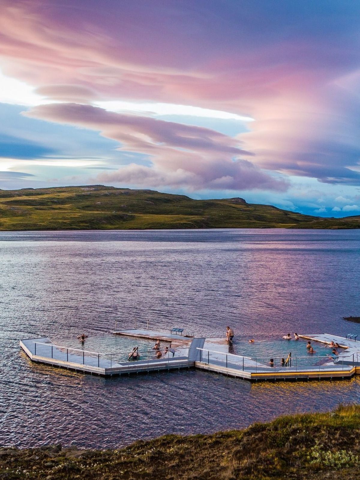 Geometric floating hot spring baths on a lake with people, surrounded by green hills under a vibrant sunset sky.