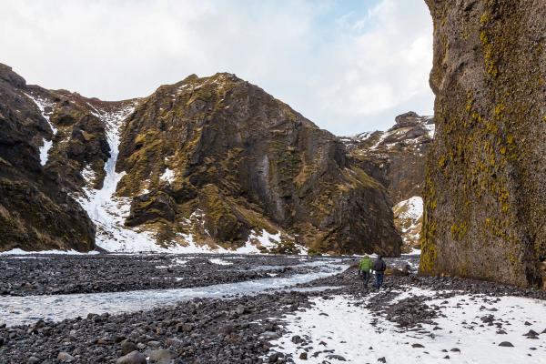 Two hikers walk along a snow-dusted, rocky riverbed through a canyon flanked by moss-covered cliffs.