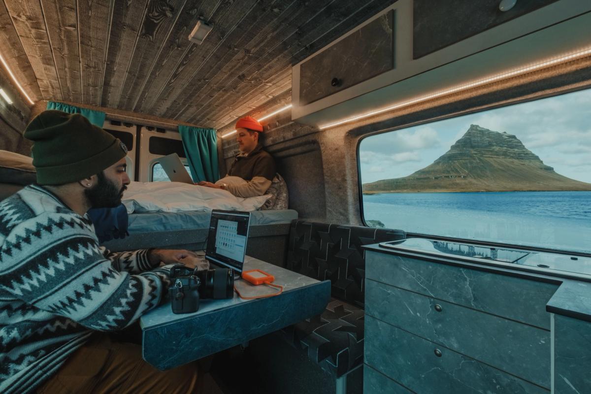 Chico sentado dentro de una camper usando el ordenador, y otro chico sentado en su cama al fondo