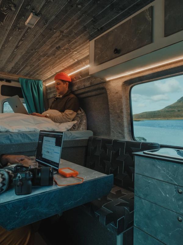 Chico sentado dentro de una camper usando el ordenador, y otro chico sentado en su cama al fondo