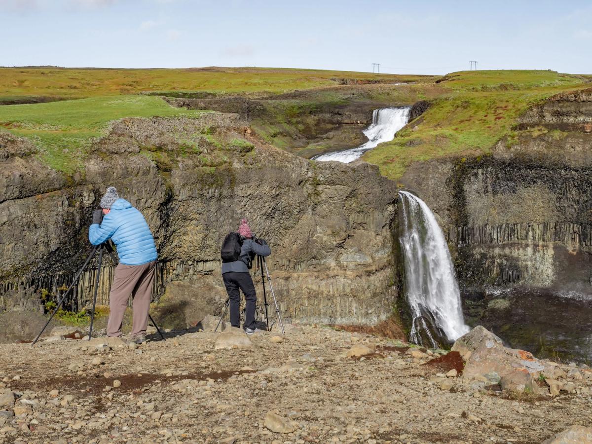 Two photographers on a rocky cliff overlook a waterfall cascading over dark columnar basalt, with a tiered waterfall on a grassy plateau in the distance.