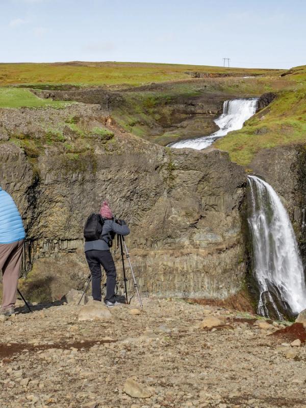 Two photographers on a rocky cliff overlook a waterfall cascading over dark columnar basalt, with a tiered waterfall on a grassy plateau in the distance.