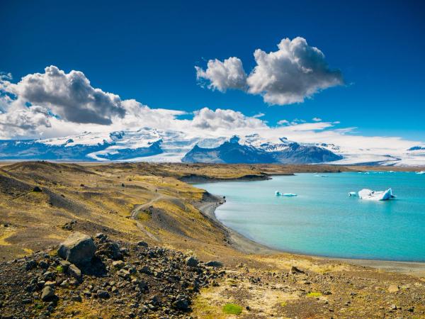 A turquoise glacial lagoon with icebergs, a rocky shore, and snow-capped mountains under a blue sky.