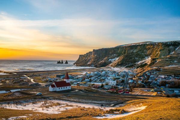 Snow-dusted coastal village with a white church, black sand beach, sea stacks, and cliffs bathed in golden light.