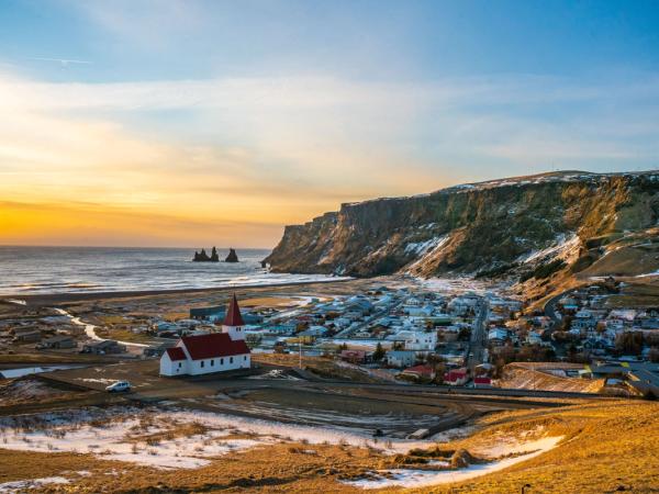 Panoramic view of a coastal village featuring a white church with a red roof, nestled between the ocean with sea stacks and rugged cliffs, under a golden sky.