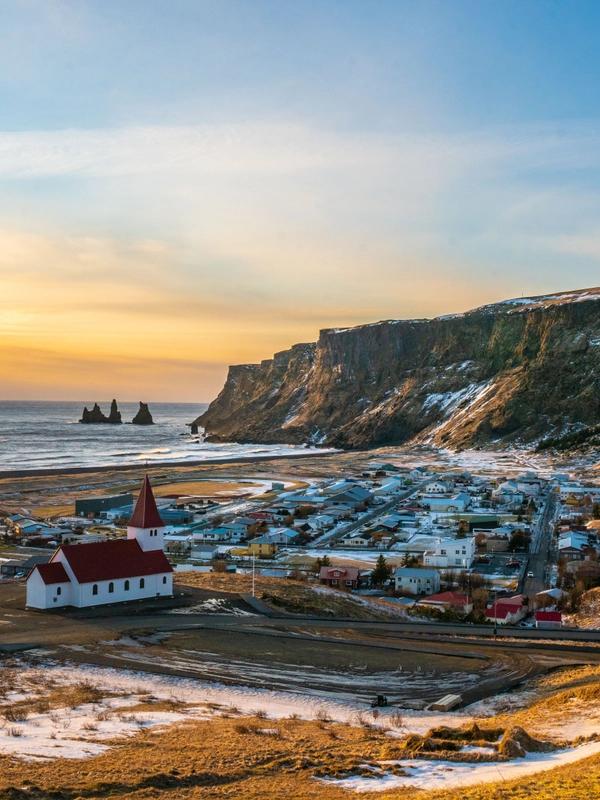 Vík í Mýrdal village with its white and red church by the sea, featuring Reynisdrangar sea stacks and cliffs under a sunset sky.