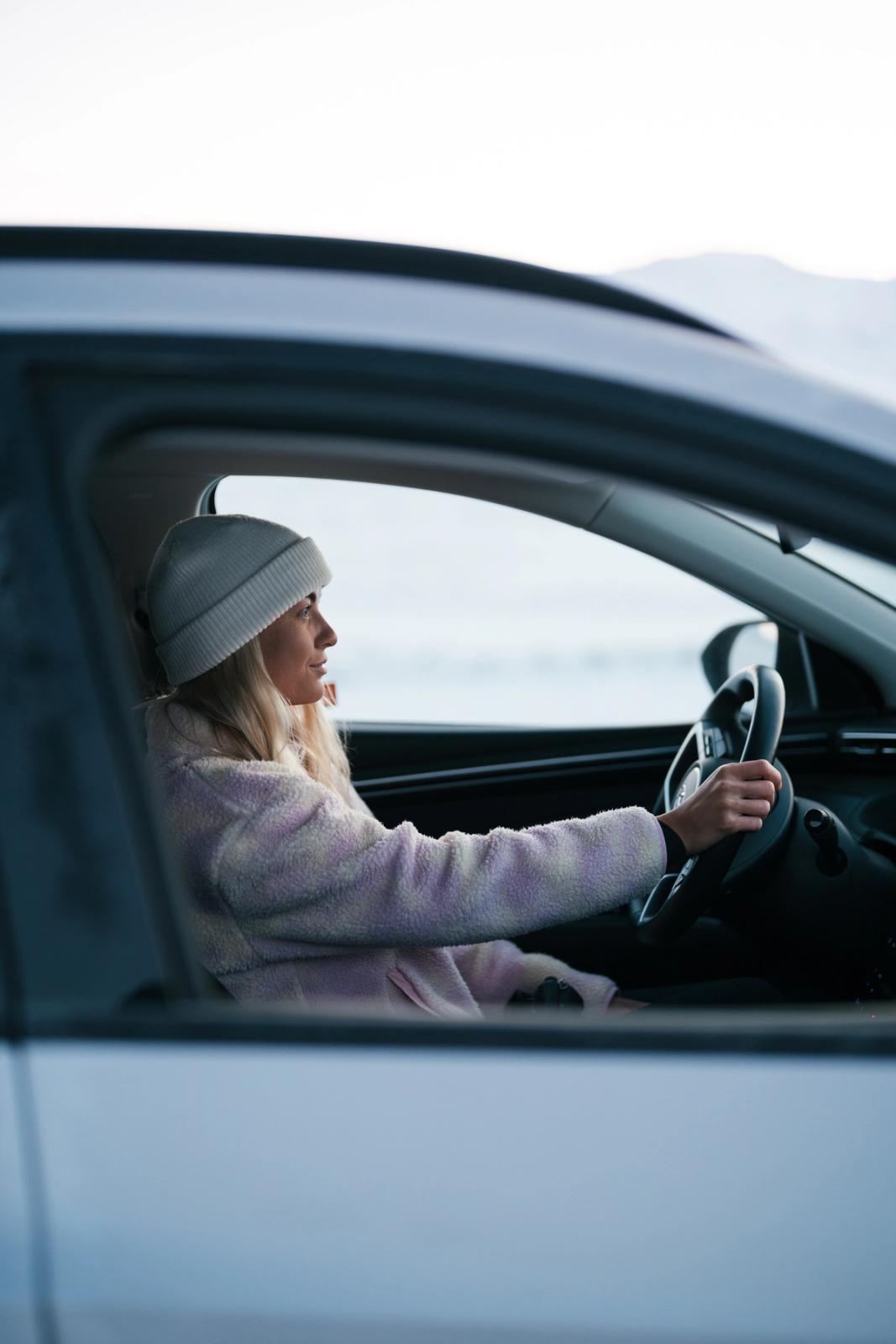 Woman in a beanie and fuzzy jacket driving a car in a winter landscape.