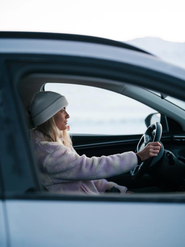 Woman in a beanie and fuzzy jacket driving a car in a winter landscape.