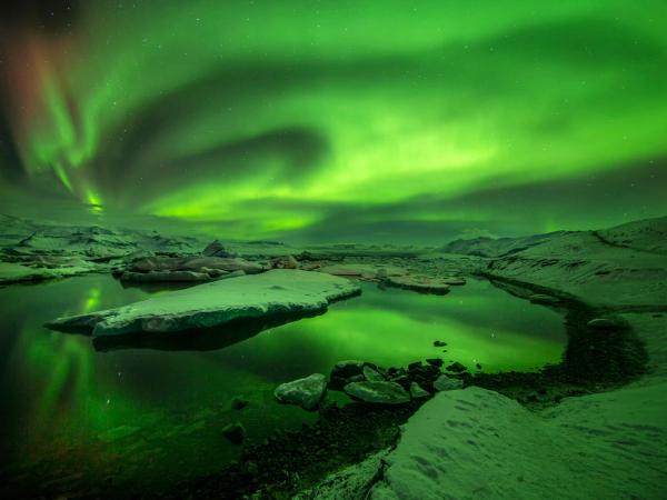 Vibrant green aurora borealis over an icy landscape with reflections in a still lake.