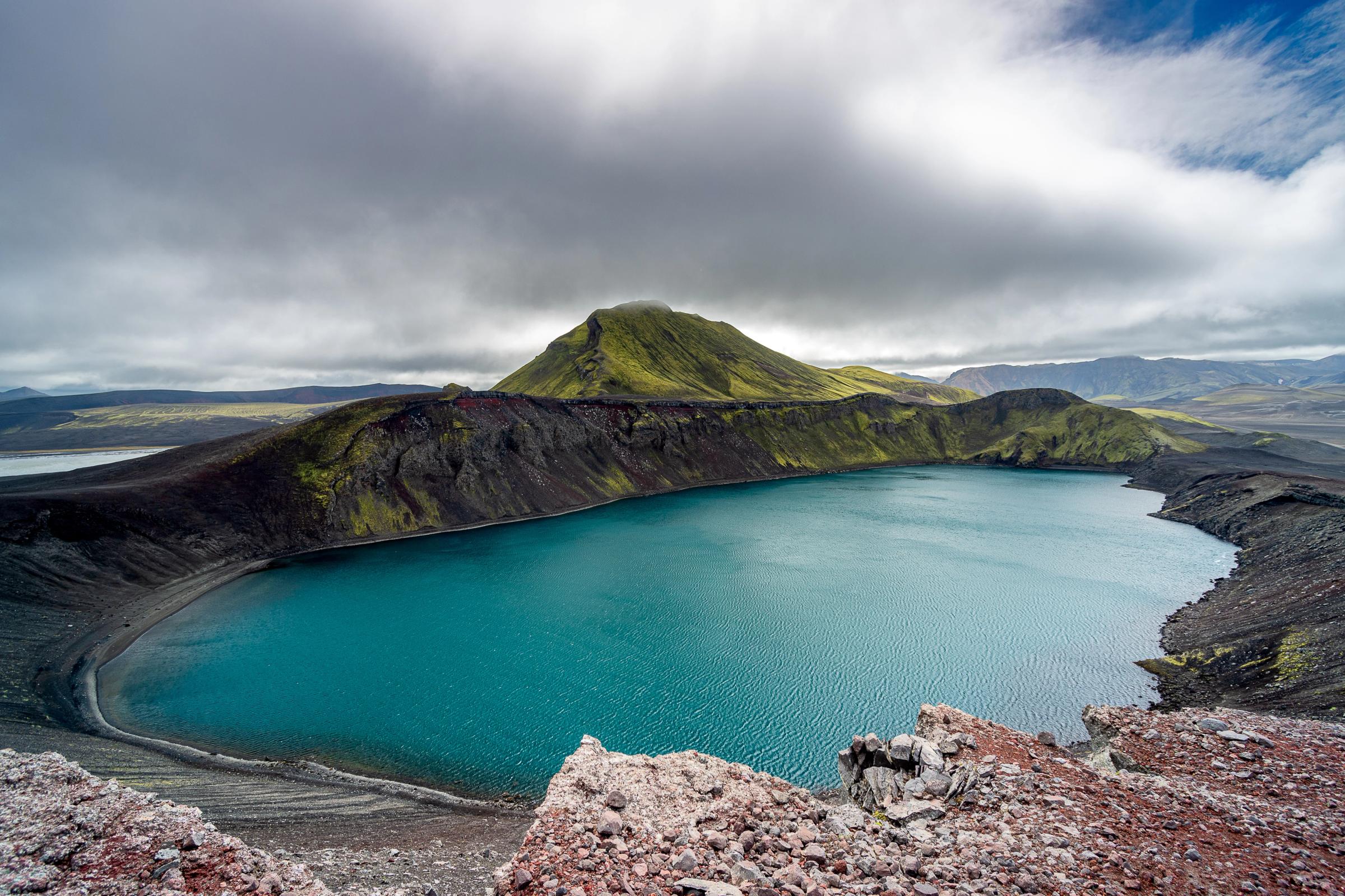 Lago de cráter turquesa en medio de un terreno volcánico oscuro y una montaña musgosa verde bajo un cielo nublado.