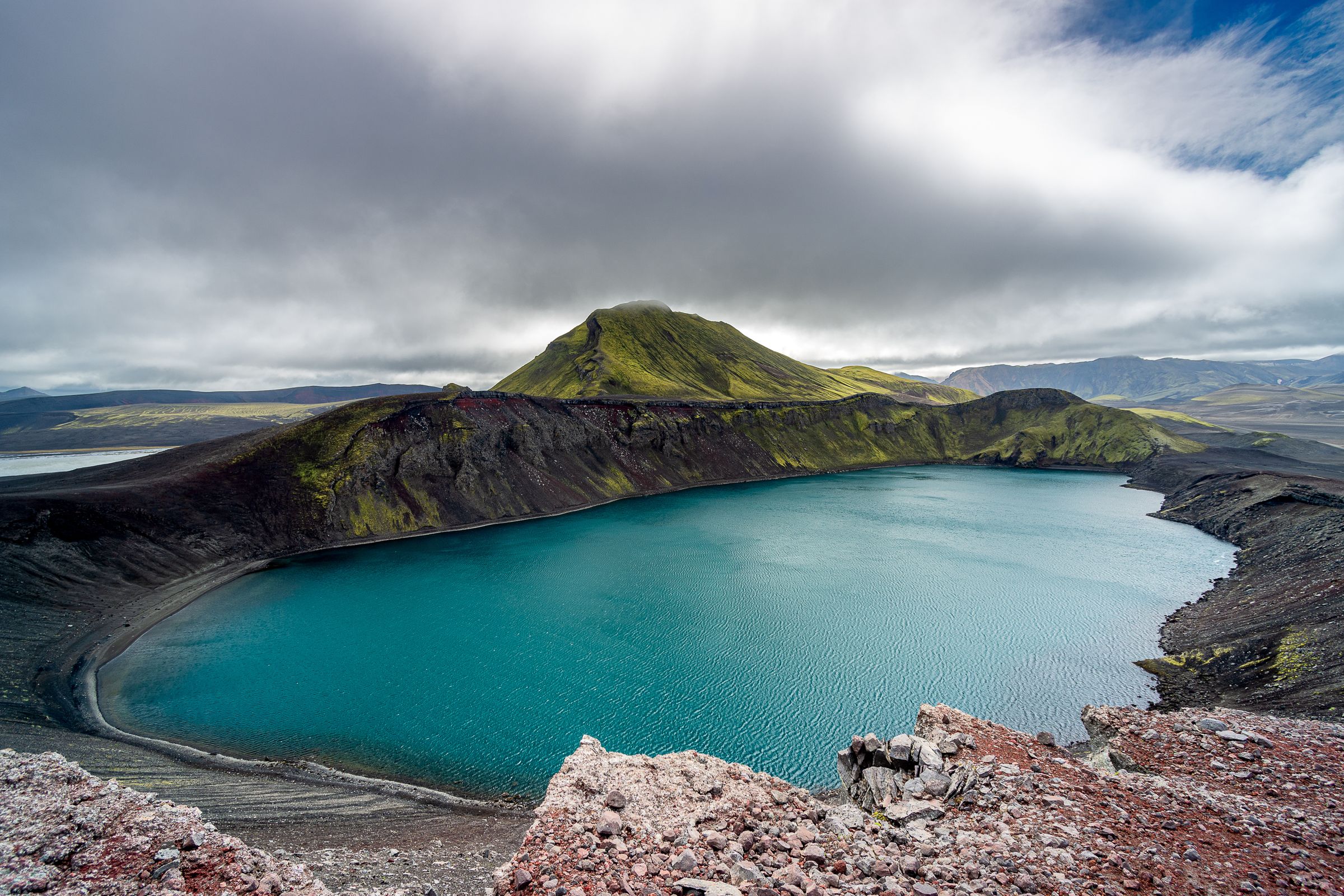 Lago de cráter turquesa en medio de un terreno volcánico oscuro y una montaña musgosa verde bajo un cielo nublado.