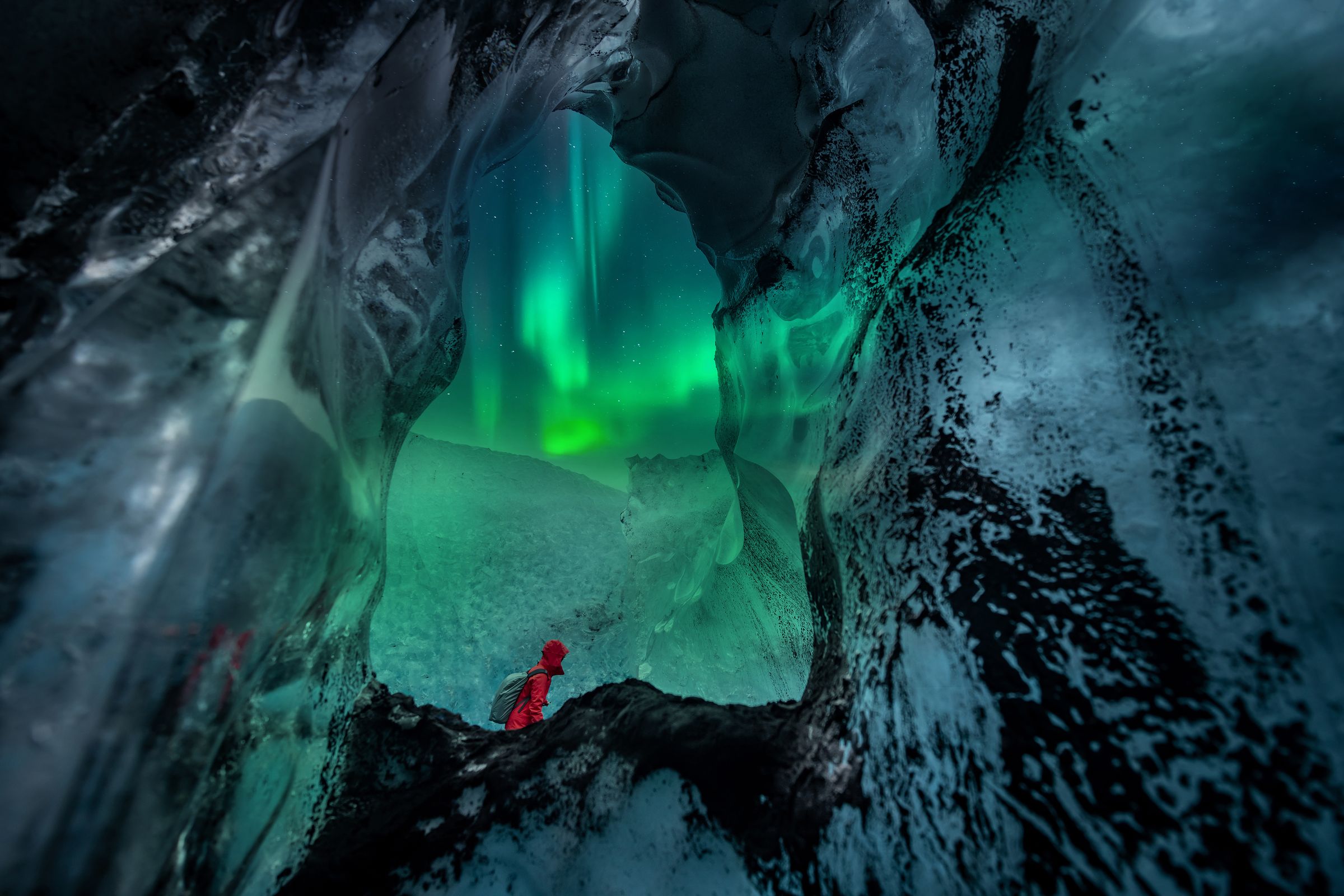 Una persona con una chaqueta roja está dentro de una cueva de hielo, mirando hacia las verdes auroras boreales.