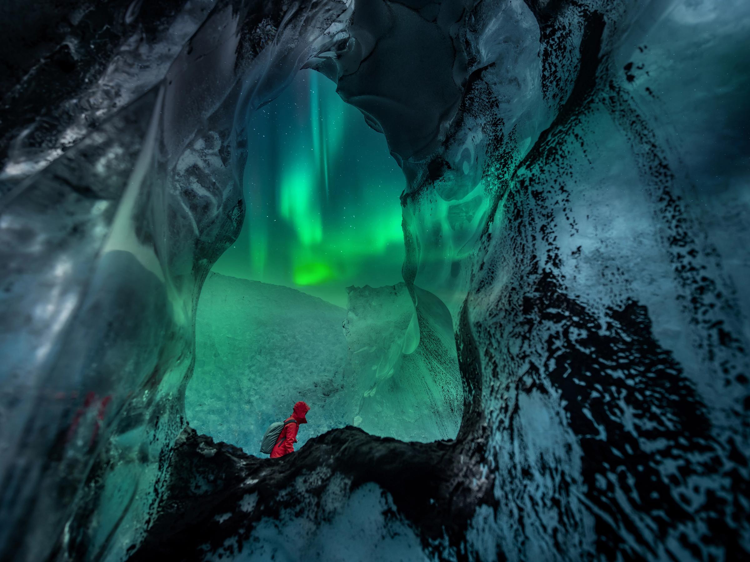 Person in red jacket inside an ice cave, looking out at the green Northern Lights.
