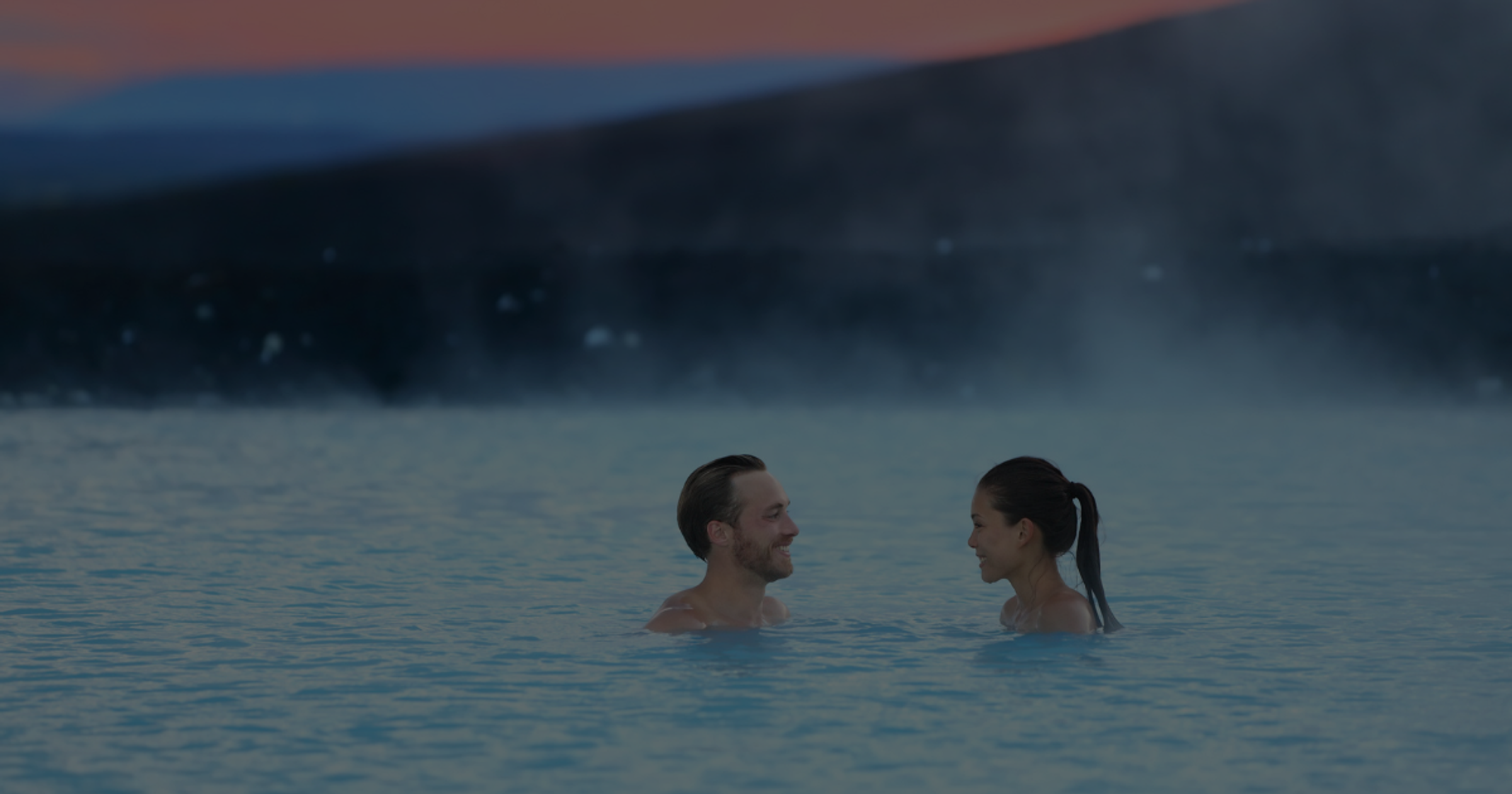 A couple smiling at each other in a steamy blue lagoon at dusk.