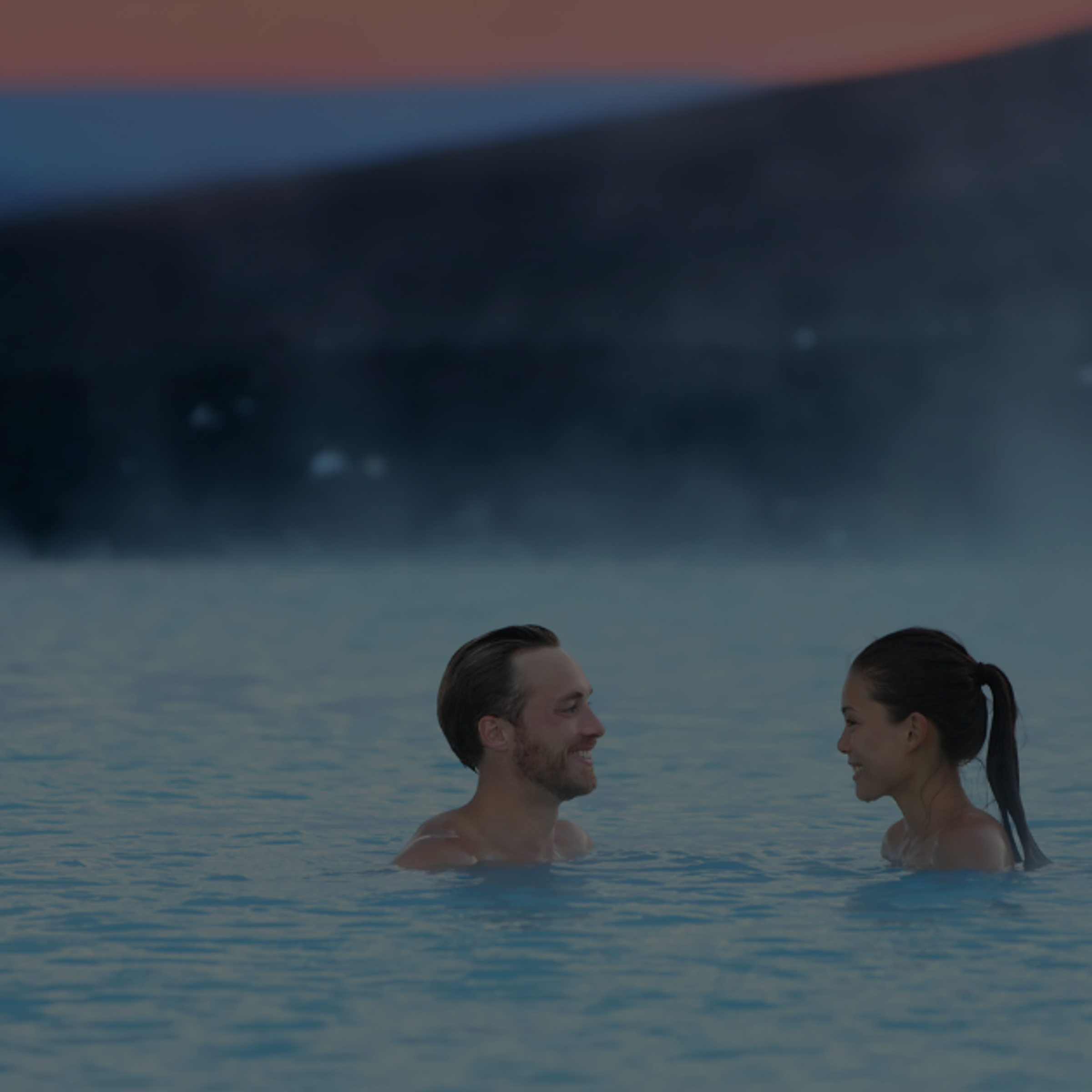A couple smiling at each other in a steamy blue lagoon at dusk.