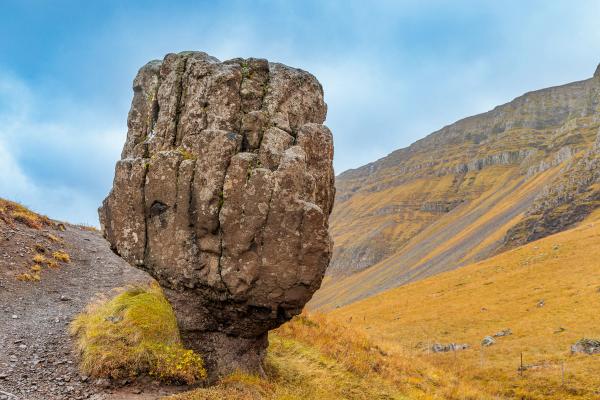 A large, rugged rock formation stands beside a dirt path, with golden-brown hills and steep mountains under a blue sky.
