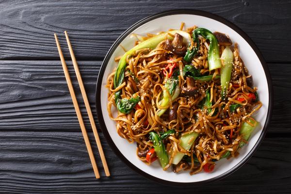 A plate of stir-fried noodles with bok choy, mushrooms, and sesame seeds, next to chopsticks.