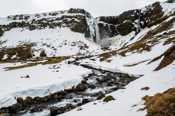 A rocky river flows through a snow-covered valley with a partially frozen waterfall cascading from cliffs in the background.