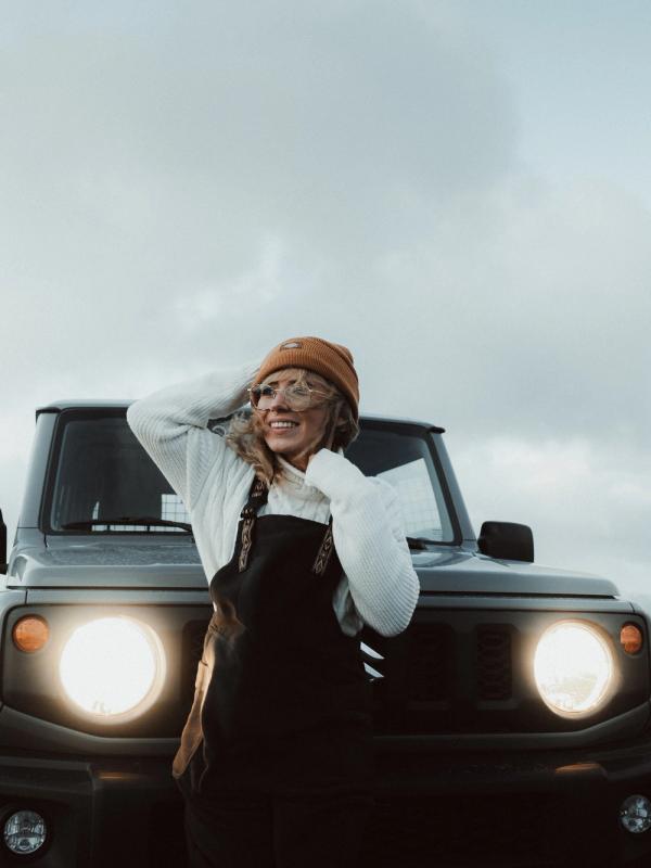 A smiling woman in a beanie and overalls stands by a gray SUV with its headlights on under a cloudy sky.