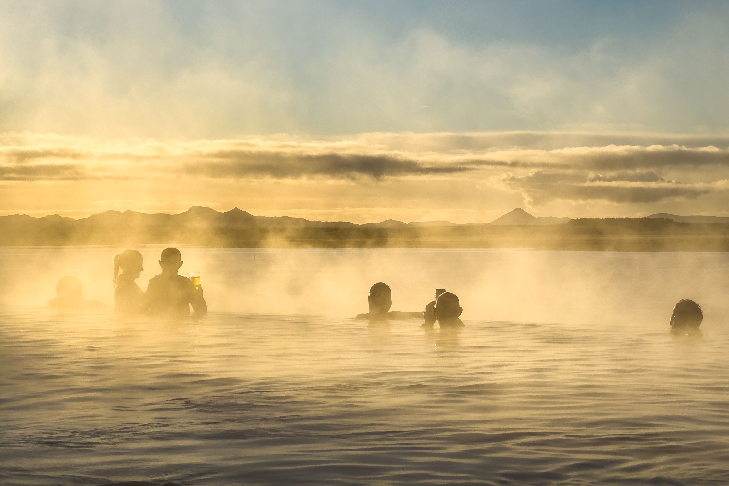 Silhouettes of people relaxing in a steaming geothermal pool at sunset in Iceland, with mountains in the distance.