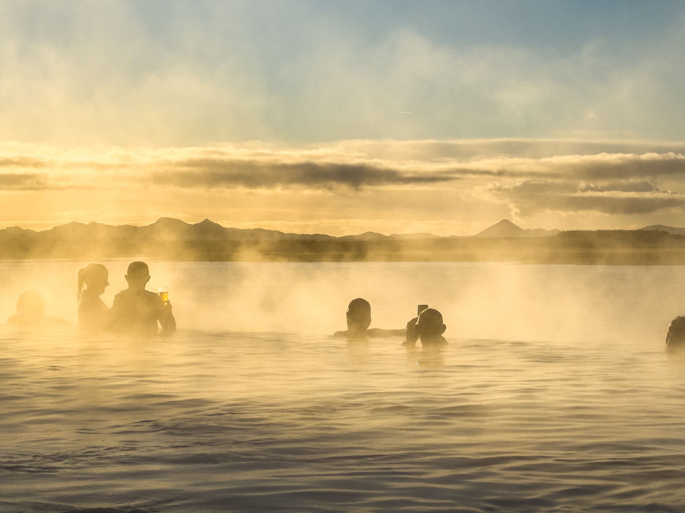 Silhouettes of people relaxing in a steaming geothermal pool at sunset in Iceland, with mountains in the distance.