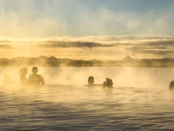 Silhouettes of people relaxing in a steaming geothermal pool at sunset in Iceland, with mountains in the distance.