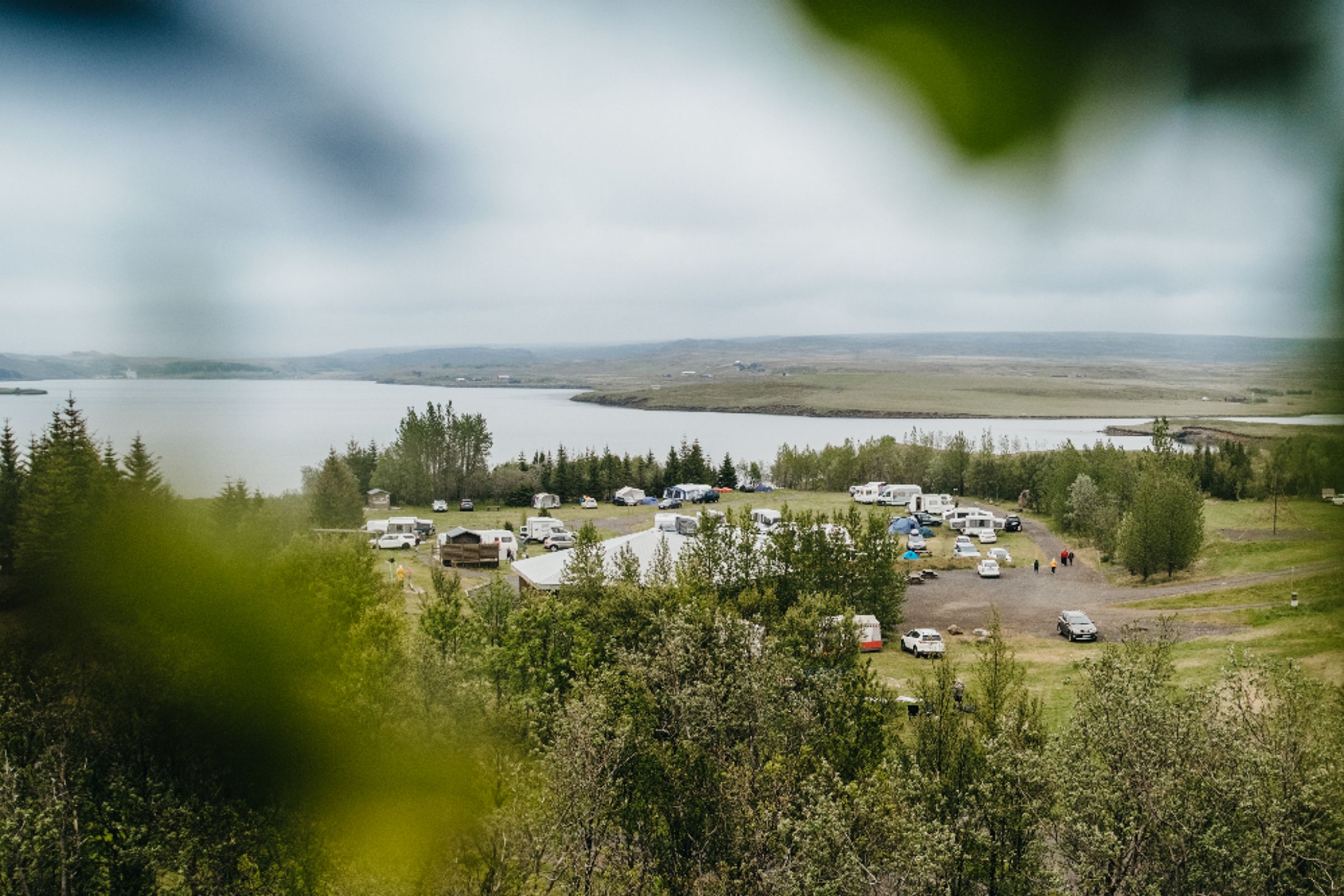A campground with RVs and tents by a lake, seen from above through green foliage on a cloudy day.