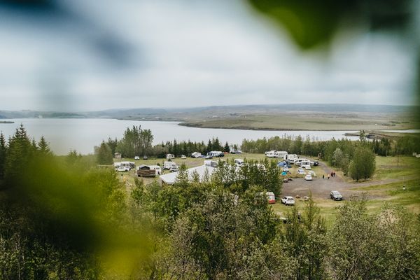 A campground with RVs and tents by a lake, seen from above through green foliage on a cloudy day.