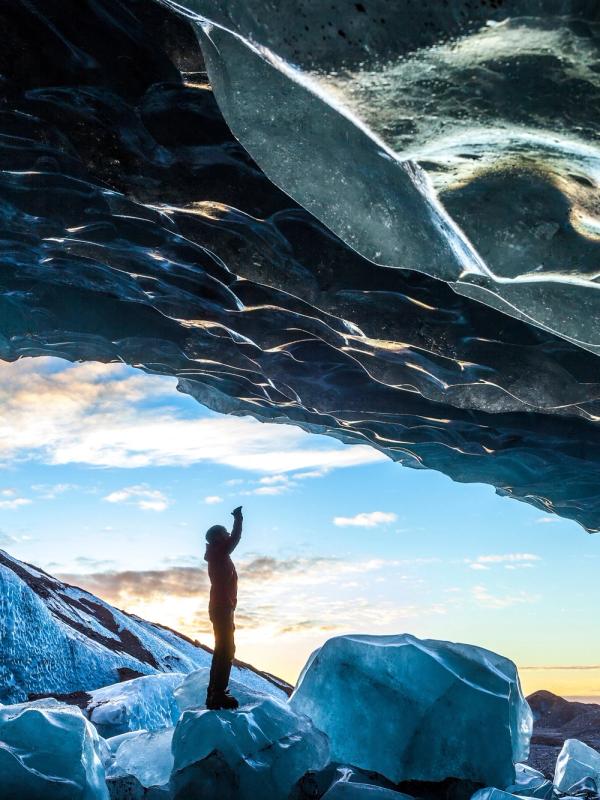 A person stands inside a large blue ice cave, pointing up at the textured icy ceiling, with a bright sky visible through the opening.