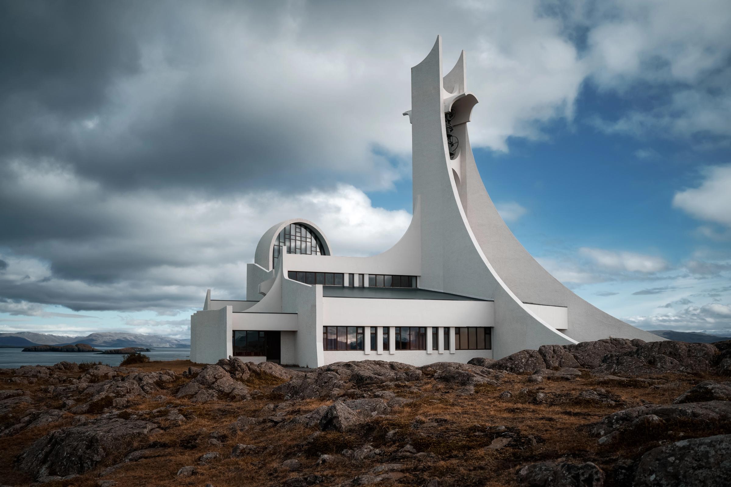A striking, white modernist church with a tall, curved spire sits on a rocky coastal landscape under a dramatic sky.